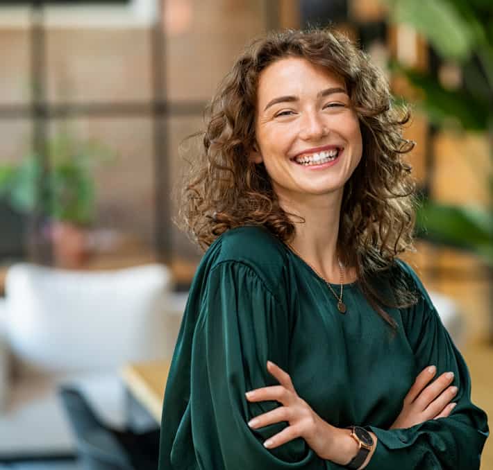 A smiling person with curly hair stands indoors, embodying a digital twin of confidence, wearing a green top and a watch, arms crossed.